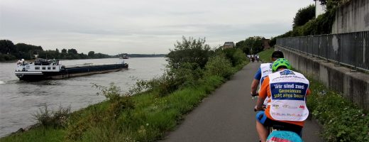 Auf dem Rhein ein Schiff, auf dem Fahrradweg daneben Teilnehmer des Tandem-Camps